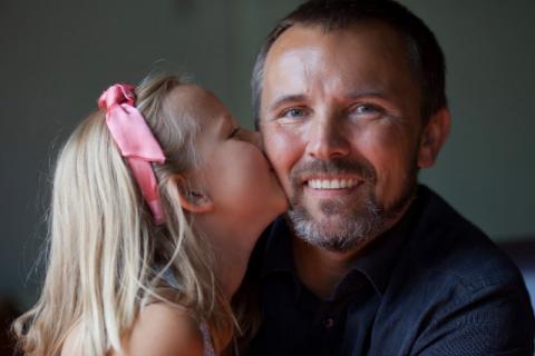 女儿头上戴着一个粉色蝴蝶结亲吻着她微笑的父亲的脸颊