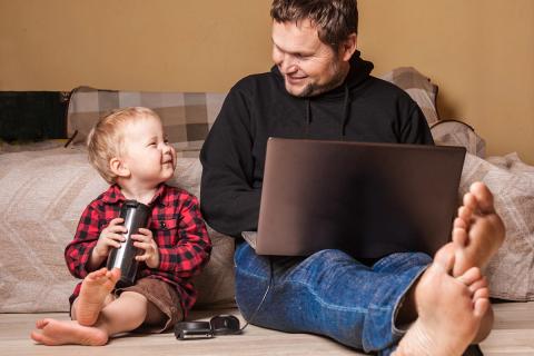 父亲在笔记本电脑上工作，年幼的儿子快乐地坐在他身边。