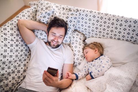 父亲一边笑一边看手机，一边躺在沙发上，旁边是正在睡觉的蹒跚学步的孩子。
