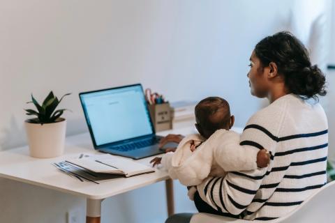 一位母亲将婴儿抱在膝上，而她在桌子上使用笔记本电脑。