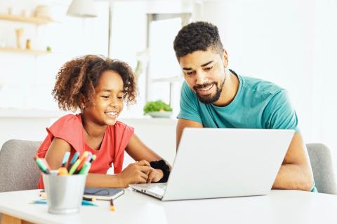 一对父女坐在书桌前，一边看着笔记本电脑一边微笑着，旁边放着一支笔。
